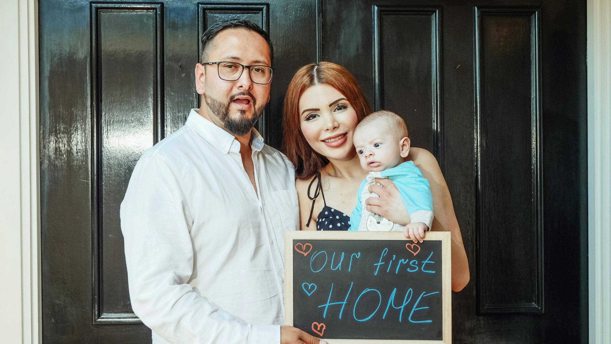 A non-resident couple posing in front of the door to a house, with the female holding a baby, and both of them holding up a sign that says our first home.