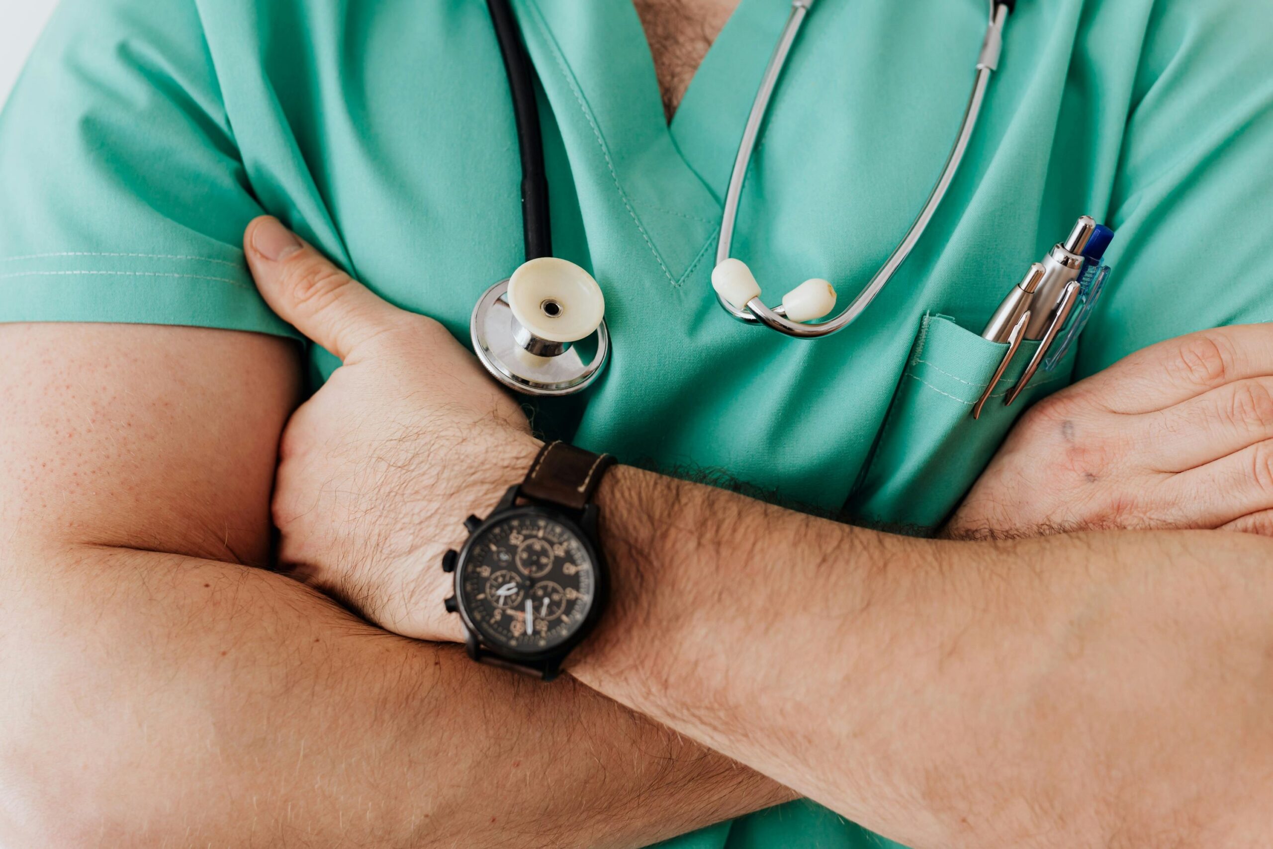 A male doctor wearing a green scrub suit and a wristwatch, standing confidently in a clinical setting.